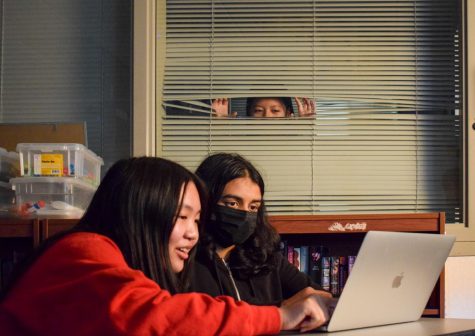 Students minding their own buisness while browsing the web together, while a mysterious figure stalks over them from behind blinds as to be surveilling them.