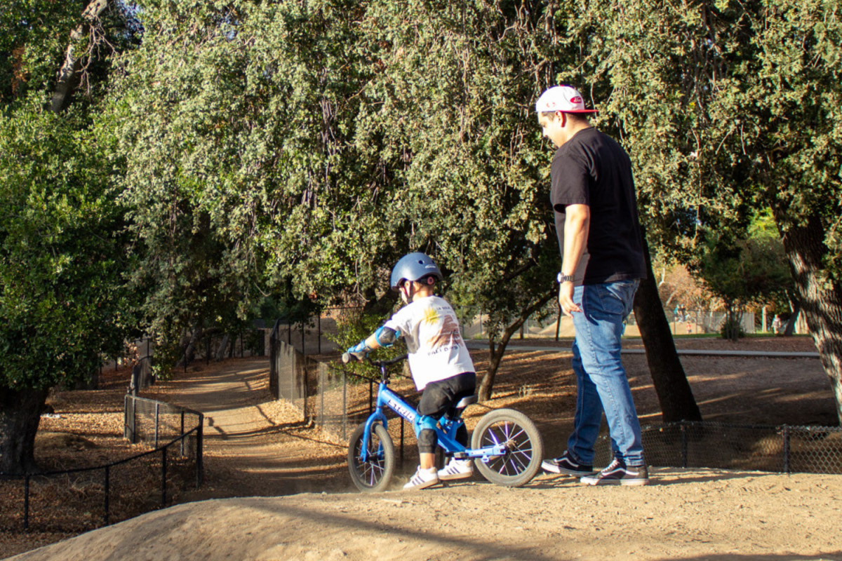 As a kid rides a bike down the hill, their dad trails closely behind.